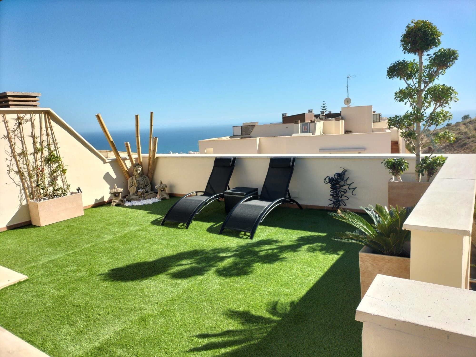 A rooftop terrace with artificial grass, two lounge chairs, a Buddha statue, and a view of the sea.