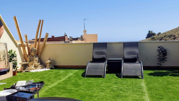 Une terrasse sur le toit, recouverte de gazon synthétique, avec deux chaises longues, une petite statue de Bouddha et une antenne parabolique.