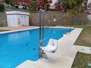 Ein Schwimmbecken mit einem Rettungsschwimmerstuhl und einem Sicherheitsschild, umgeben von einer Steinmauer und einem kleinen Gebäude mit rotem Dach.