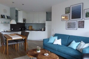 A modern living room with a blue sofa, wooden coffee table, and a dining area with a table and chairs. - Holiday apartment 14 in Villa Meeresblau (Cuxhaven)