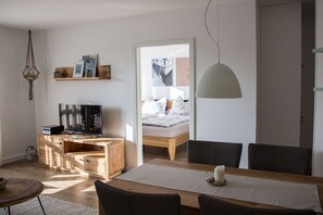 A modern living room with a wooden TV stand, a dining table with chairs, a hanging light fixture, and a bed with a white headboard in the background. - Holiday apartment 14 in Villa Meeresblau (Cuxhaven)