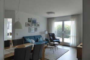 A modern living room with a blue sofa, a dining table with chairs, a coffee table, and a large window with curtains. - Holiday apartment 14 in Villa Meeresblau (Cuxhaven)