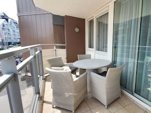 Ein Balkon mit einem runden Tisch und Korbstühlen, ein Fenster mit Vorhängen und Blick auf Gebäude.