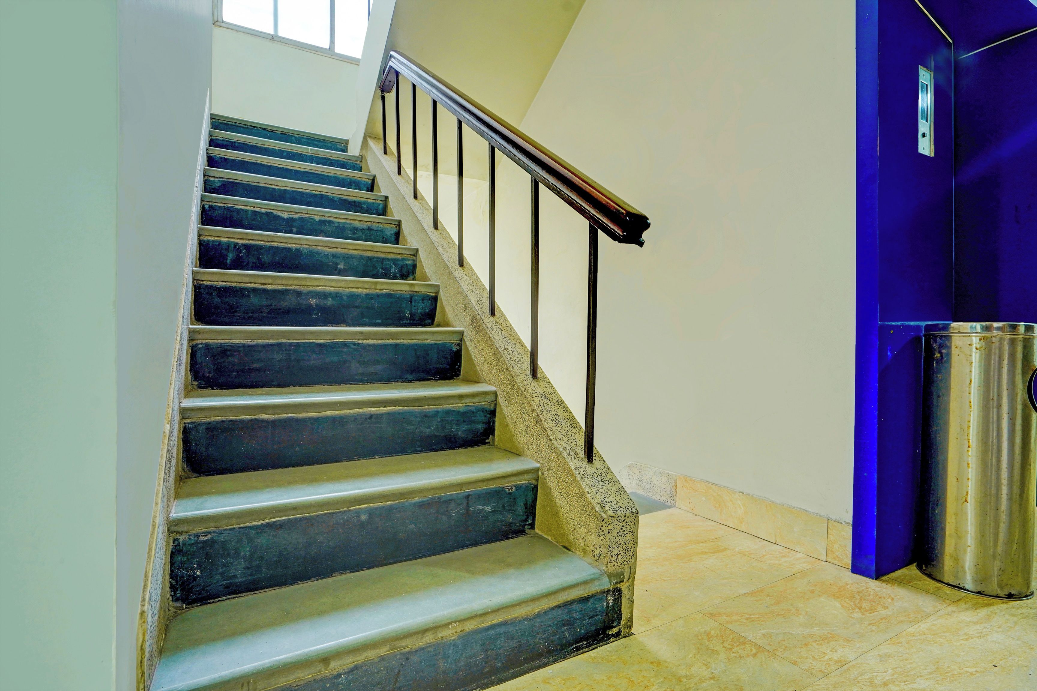 A staircase with a metal handrail, a trash bin, and a blue door.