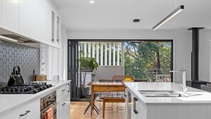 A modern kitchen with white cabinetry, a gas stove, and a dining area with a wooden table and chairs. - Bangalay on Tallwood (Mollymook Beach)