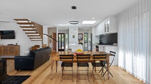 A modern kitchen with a central island, wooden flooring, and a dining area with a table and chairs. - Bangalay on Tallwood (Mollymook Beach)