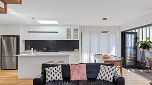 A modern kitchen with a central island, stainless steel appliances, and a dining area with a view. - Bangalay on Tallwood (Mollymook Beach)