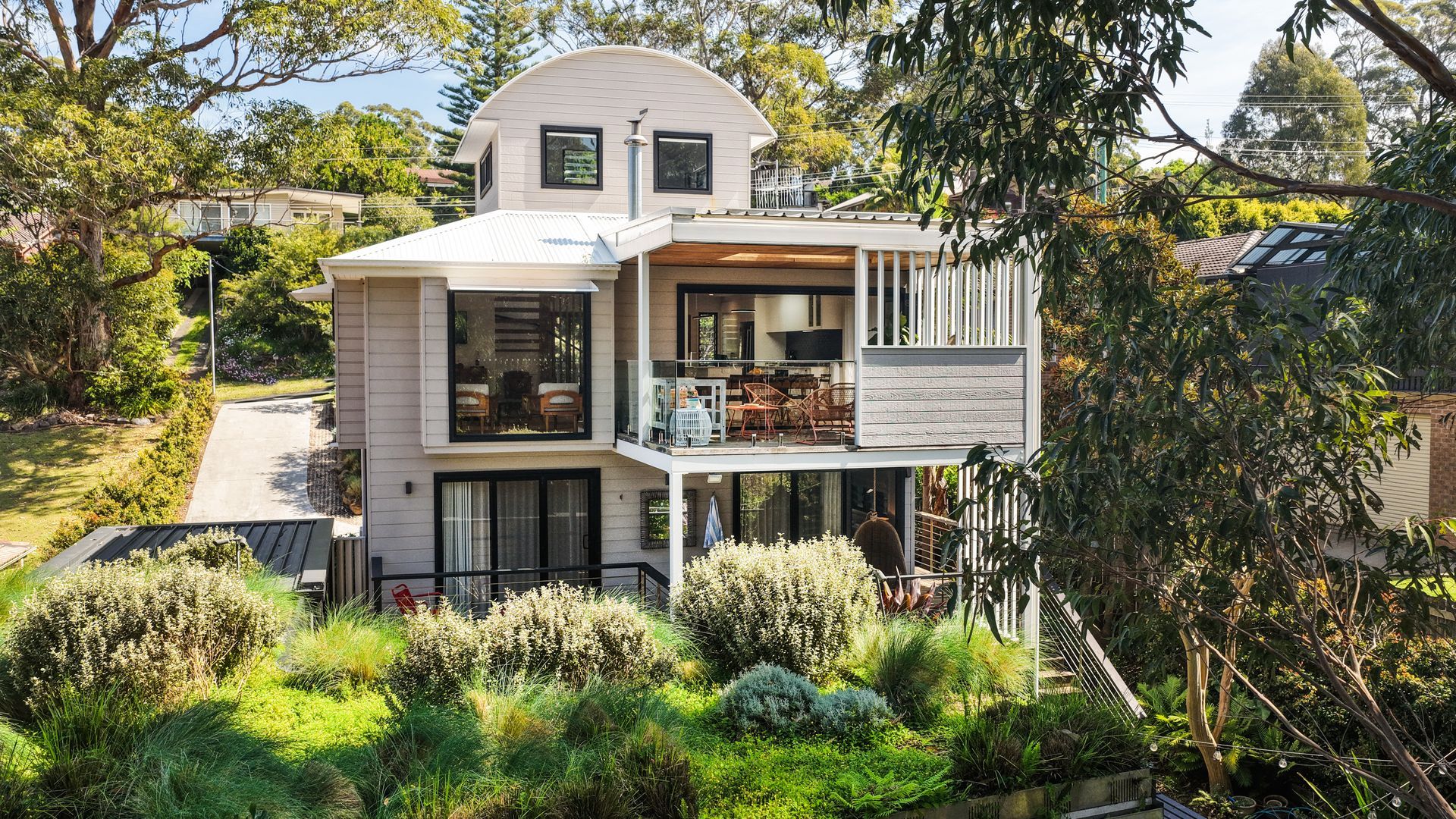 A modern two-story house with a balcony, surrounded by lush greenery.