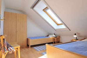 Une petite pièce bien éclairée, avec un plafond en pente, un lit simple, une chaise, une armoire et un bureau avec une lampe.
