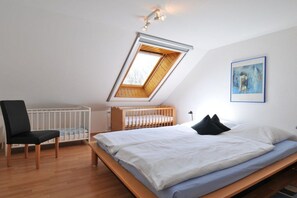 A bedroom with a sloped ceiling, a crib, a chair, and a bed with pillows. - Holiday apartment with 1 bedroom (Carolinensiel)