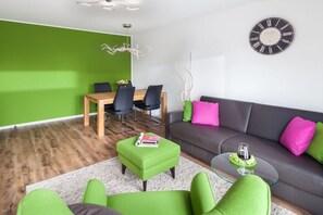 A modern living room with a green wall, a dining area with chairs and a table, a grey sofa with colorful cushions, and a wall clock. - Apartment in Juist with swimming pool (Juist)
