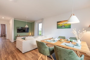 A modern living room with a sofa, dining table, and chairs. There is a television mounted on the wall and a painting above the table. - Holiday apartment with 1 bedroom (Lohme OT Ranzow)