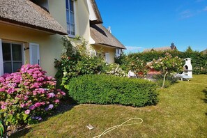 Ein traditionelles Haus mit einem Reetdach, ein gepflegter Garten mit rosa Blumen und im Hintergrund ein weißer Glockenturm.
