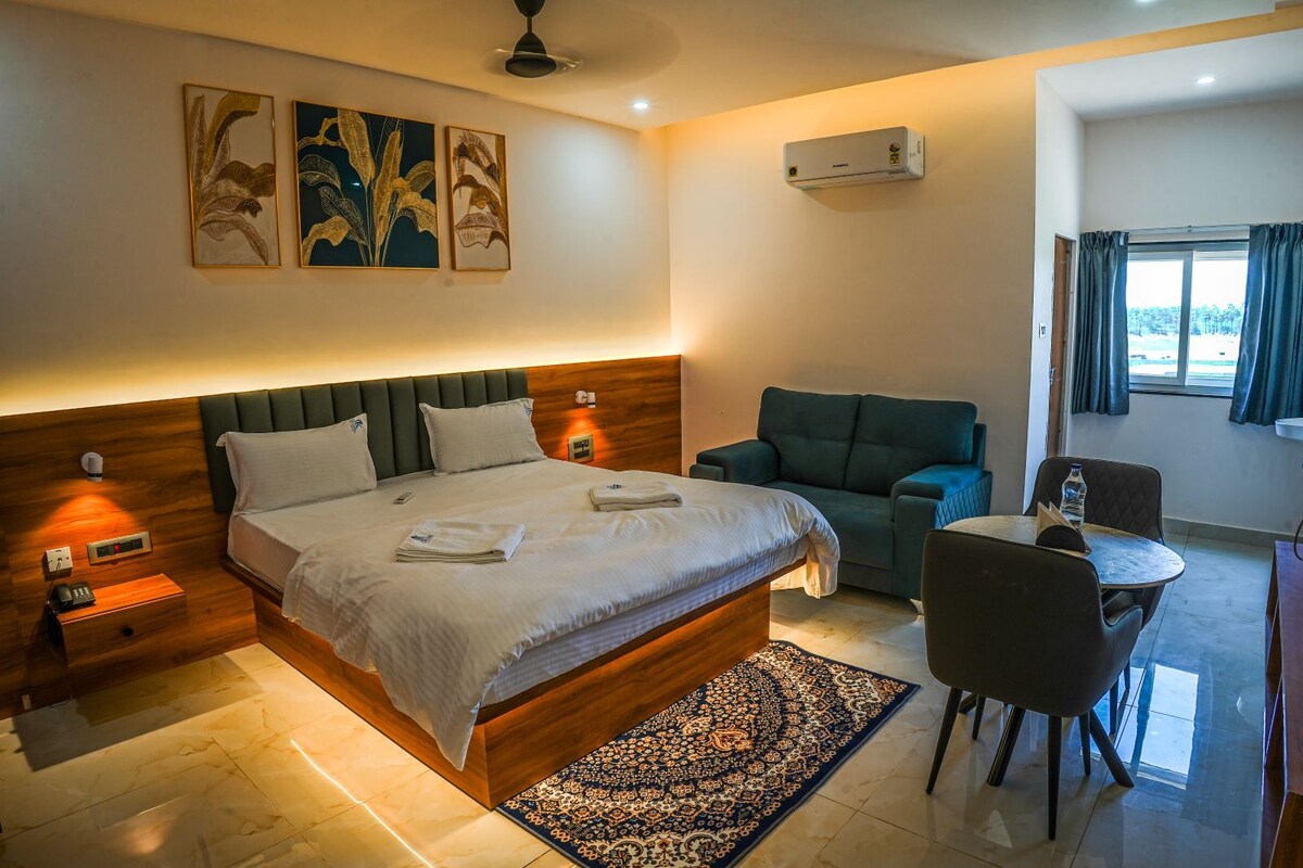 A hotel room with a bed, bedside table, lamp, chair, table, and a window with curtains.