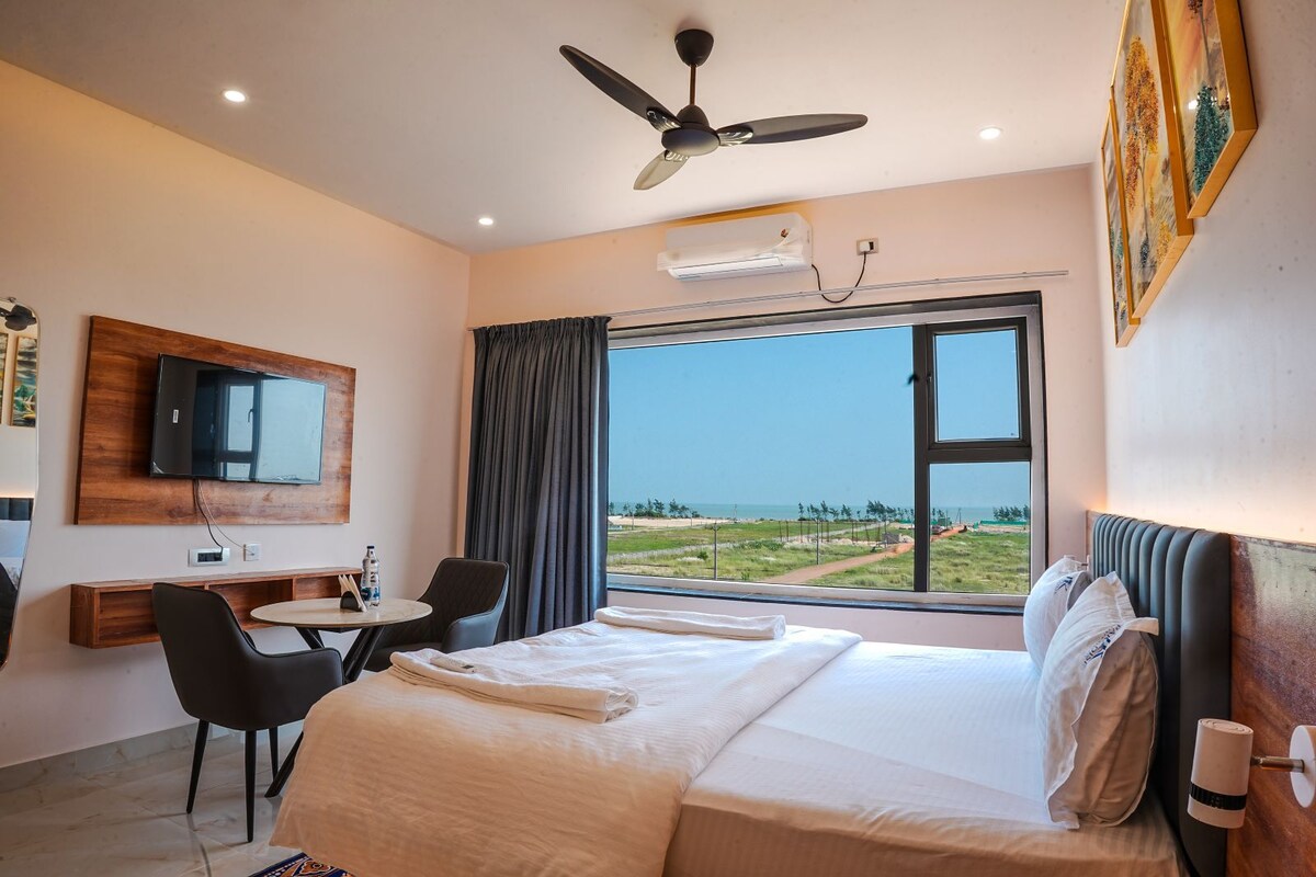 A hotel room with a bed, a desk, a chair, a television, and a window with a view of the beach.