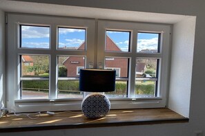 Une pièce avec une fenêtre donnant sur un quartier résidentiel, une table ronde avec une lampe et une prise électrique sur le rebord de la fenêtre.