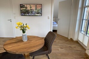 Een kamer met een ronde houten tafel, een enkele stoel, een vaas met gele bloemen en een aan de muur hangend schilderij.