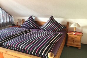 A bed with striped bedding and two pillows, a wooden nightstand with a lamp, and a window with curtains. - Holiday apartment Höflich "Heide 1" (Bad Zwischenahn)