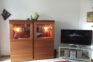 A wooden cabinet with glass doors containing a lit fireplace, a television on a stand, and a small shelf with electronic devices. - Holiday apartment Höflich "Heide 1" (Bad Zwischenahn)