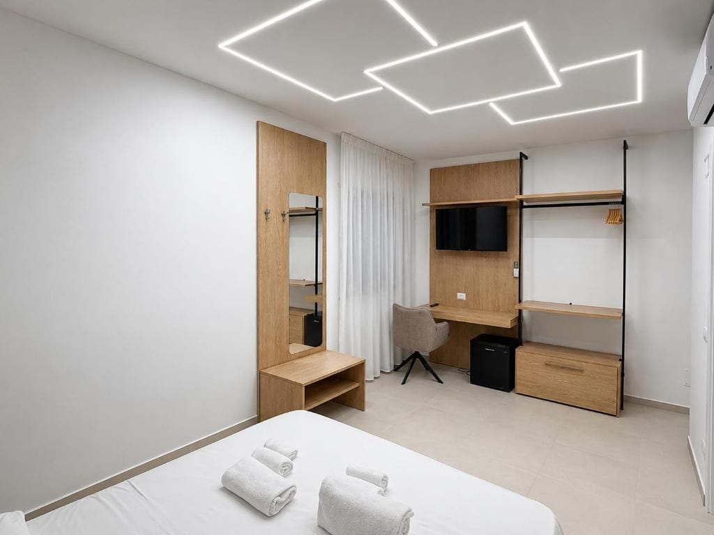 A modern hotel room with a bed, a desk, a chair, a television, and a wardrobe.