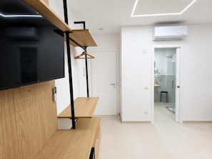 A modern interior with a flat-screen TV mounted on a wooden cabinet, a hallway leading to a bathroom, and an air conditioning unit on the wall. - Emma's Rooms (Tropea)