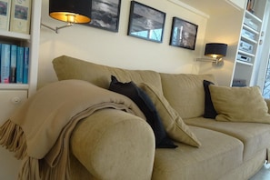 A beige sofa with a throw blanket and pillows, a bookshelf with books, and wall-mounted lamps. - Holiday apartment in Sylt with 1 bedroom (Wenningstedt-Braderup)