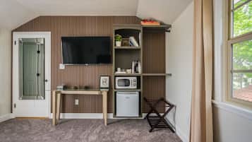 Quarto compacto com televisão integrada, micro-ondas, mini-frigorífico, estante de madeira e uma cadeira.