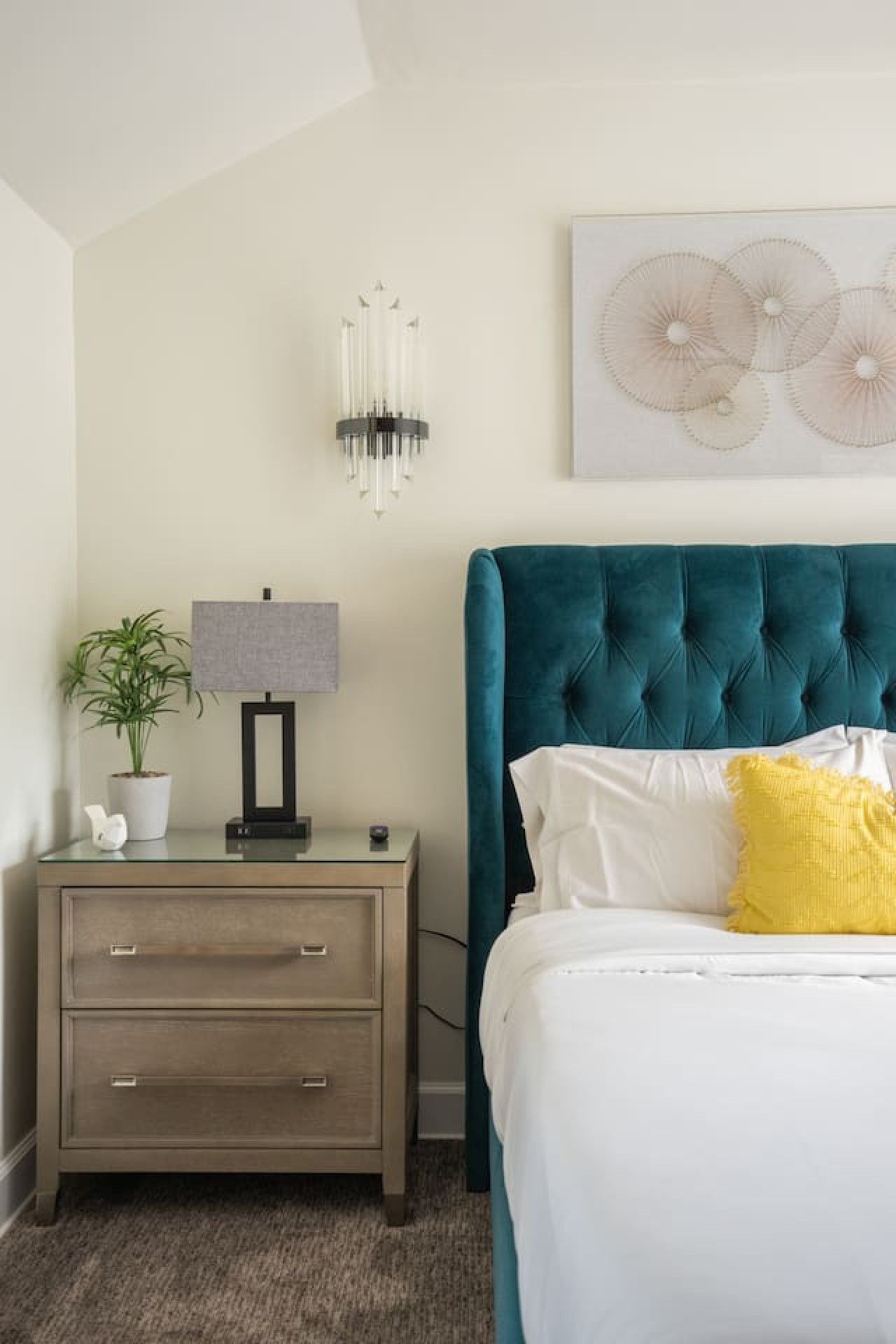 A bedroom with a bed, a nightstand, a lamp, a potted plant, and a wall art piece.
