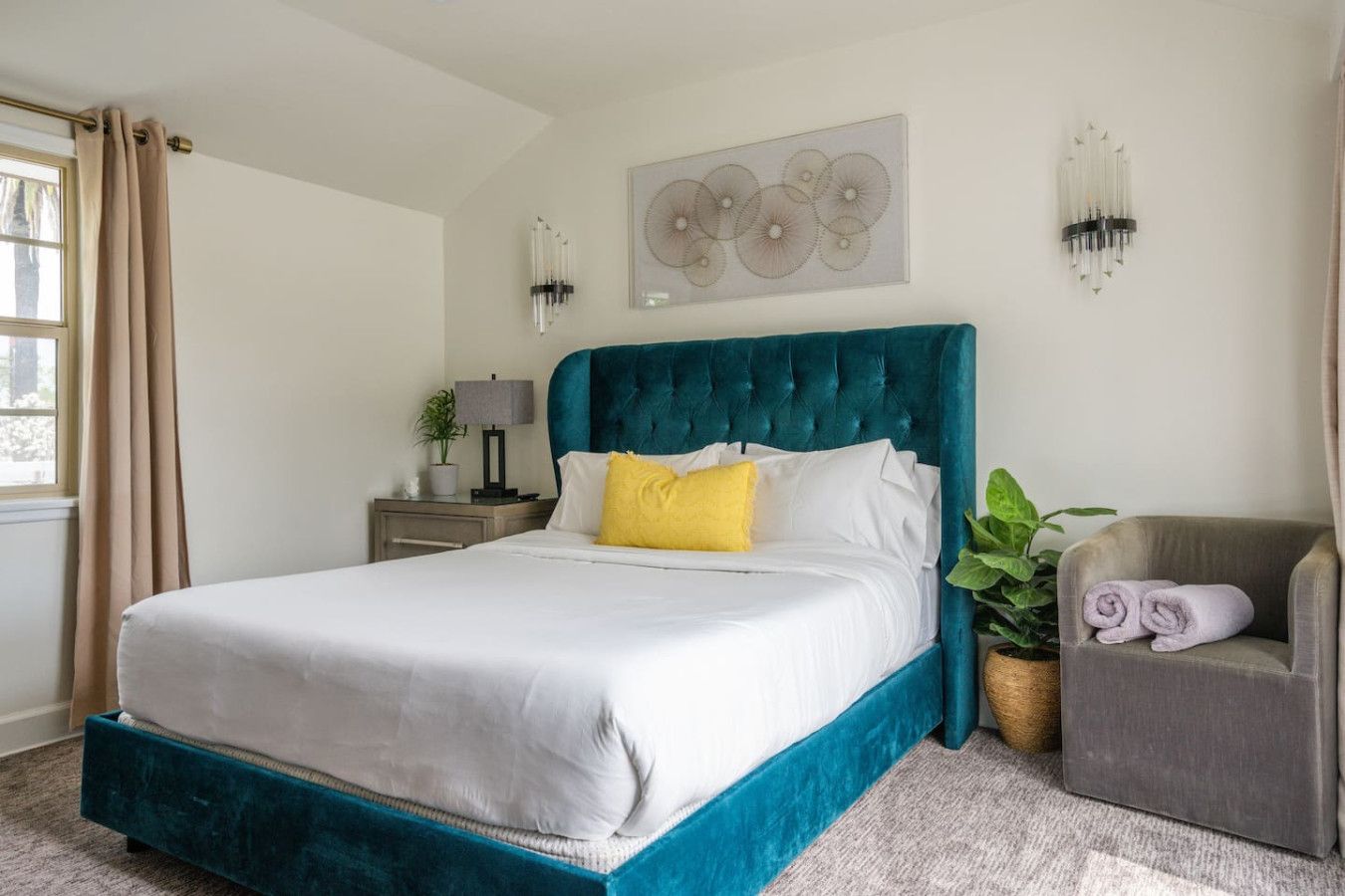 Quarto de hotel com uma cama grande, mesa de cabeceira com abajur, cadeira com toalha e uma obra de arte na parede.