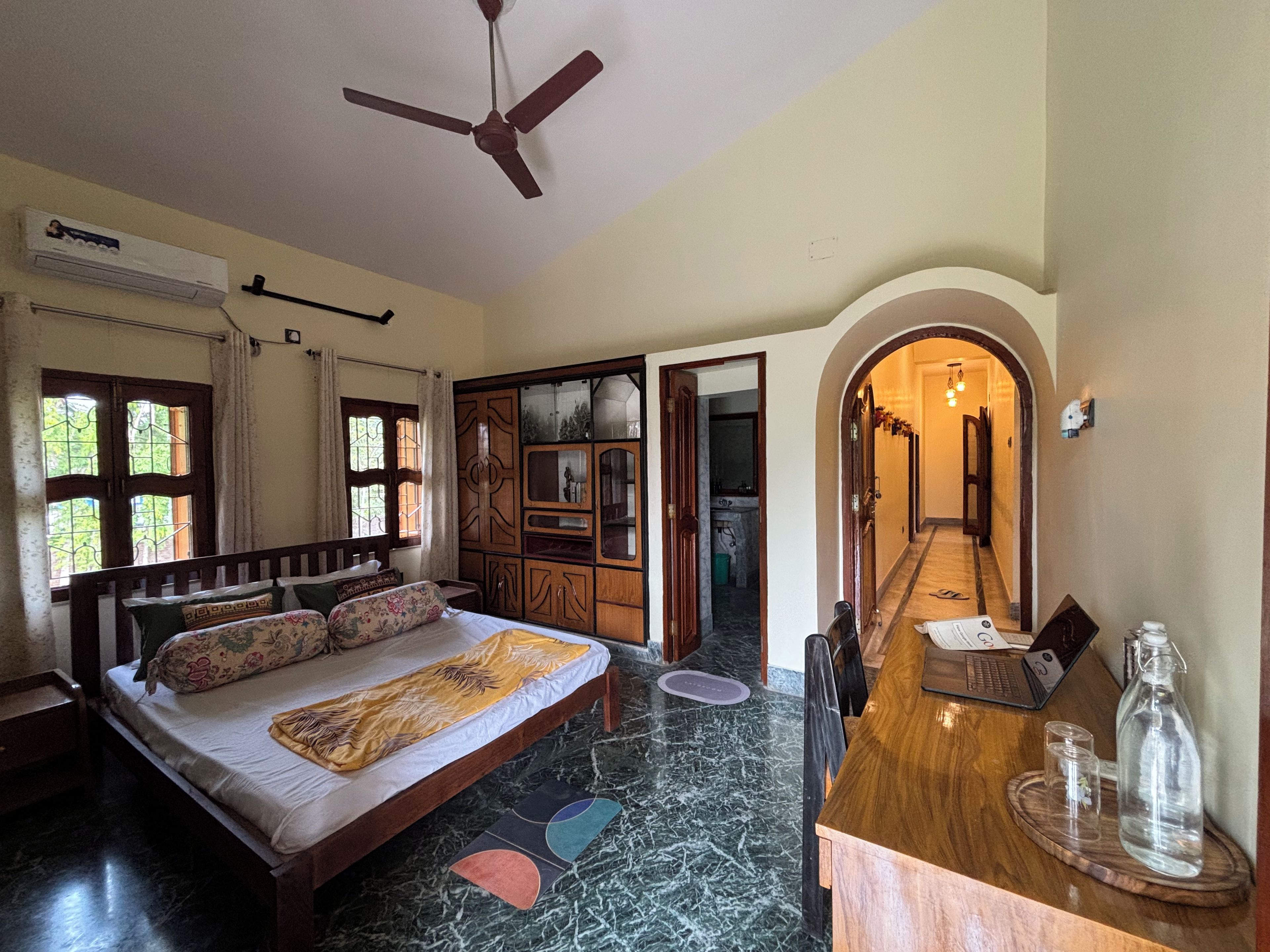 A bedroom with a bed, a ceiling fan, an air conditioning unit, a wooden wardrobe, and a bathroom visible through an open door.