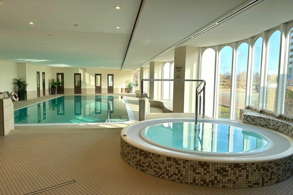 A large indoor swimming pool with a hot tub, surrounded by tiled walls and large windows.