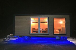 A modern mobile home with two large windows, a visible living area with furniture, and a blue-lit foundation. - Bruns holiday apartment (Ihlow)