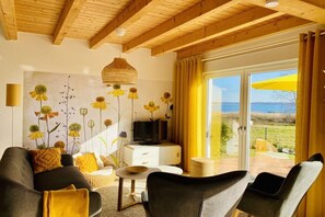 A living room with a sofa, a coffee table, and a television on a white cabinet. There is a large window with curtains and a view of the outdoors. - Holiday home with 3 bedrooms (Plau am See)