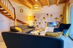 A living room with a sofa, armchair, coffee table, and a TV. There's a staircase with wooden railings and a wall-mounted lamp. - Holiday home with 3 bedrooms (Plau am See)