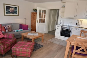 A living room with a sofa, ottoman, and coffee table, a kitchen with white cabinets and appliances, and a door leading to another room. - Holiday apartment in Sylt with 1 bedroom (Wenningstedt)