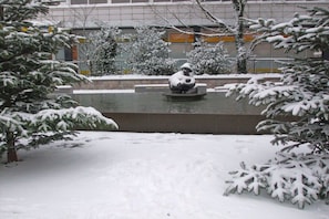 Ein verschneiter Außenbereich mit einem Brunnen und schneebedeckten Bäumen.