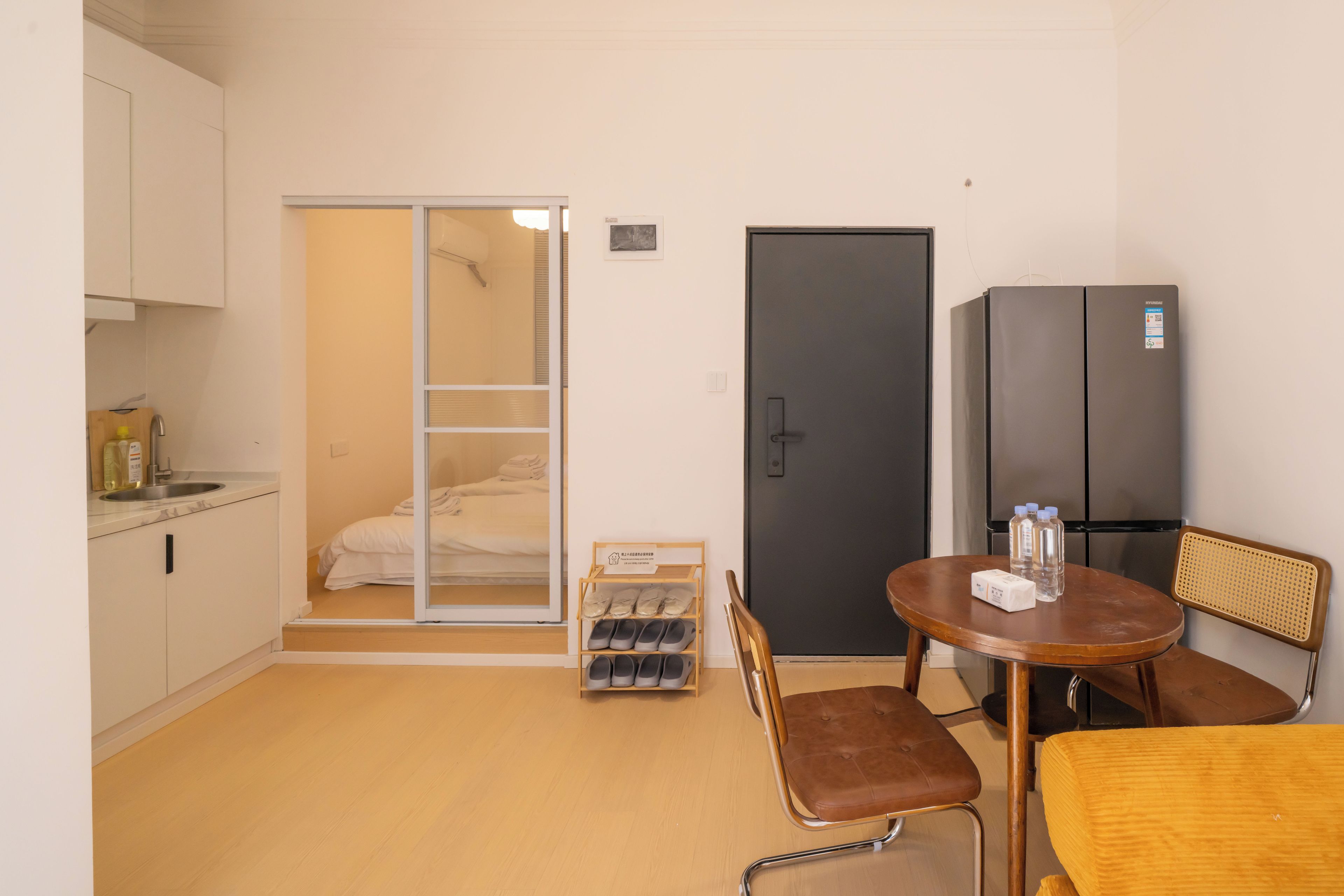A compact hotel room with a kitchenette, a bed with a sliding door closet, a refrigerator, a dining table with chairs, and a small shoe rack.