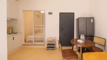 A compact hotel room with a kitchenette, a bed with a sliding door closet, a refrigerator, a dining table with chairs, and a small shoe rack.