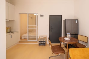 A compact hotel room with a kitchenette, a bed with a sliding door closet, a refrigerator, a dining table with chairs, and a small shoe rack. - Book&Bed-People's Sq. Changzheng Hosp. (Huangpu District)