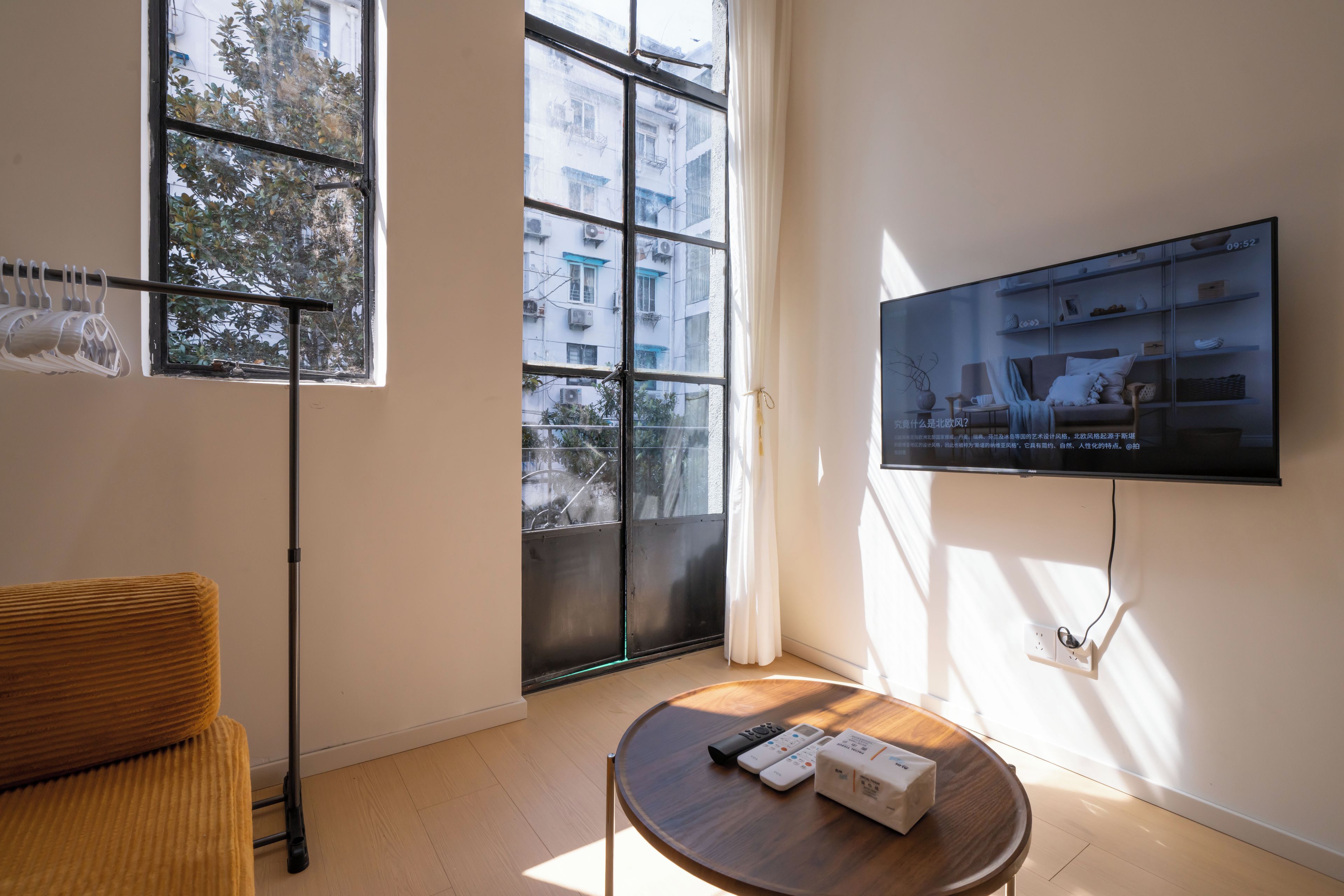 Ein modernes Wohnzimmer mit einem flachbildfernseher an der Wand, einem Couchtisch mit Fernbedienungen und einer Schachtel, einer Couch und einem Fenster mit Blick auf Gebäude und Bäume.