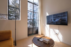 Uma sala de estar moderna com televisão de tela plana na parede, uma mesa de centro de madeira com controles remotos e uma caixa, um sofá e uma janela com vista para prédios e árvores.