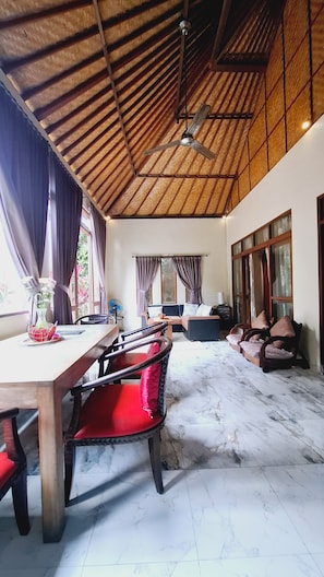 A spacious room with a high ceiling, a dining area with a wooden table and red chairs, and a seating area with sofas and a television. - Gajah Biru Ubud (Ubud,Gianyar)