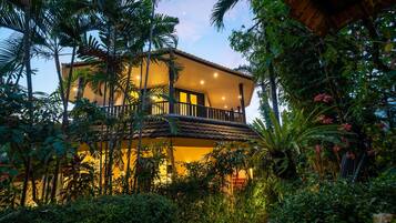 A two-story building with a balcony, surrounded by lush greenery and a thatched roof structure.