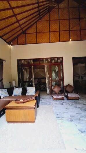 A spacious living area with a high ceiling, a sofa set, a coffee table, and a view through a large window.