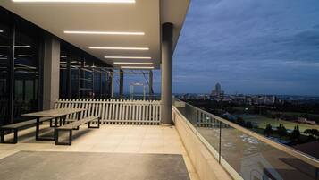 Een balkon buiten met uitzicht op een stadsbeeld, met een hekwerk, bankjes en een heldere lucht.