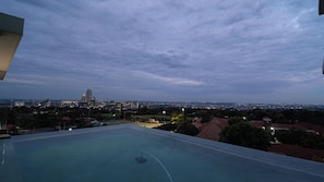 A rooftop pool with a view of a city skyline at dusk. - The Illovo Residence | Luxury 1-Bedroom Apartment (Sandton)