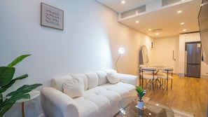 A modern living room with a sofa, a dining table with chairs, a refrigerator, and a potted plant. - The Illovo Residence | Luxury 1-Bedroom Apartment (Sandton)