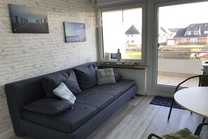 A modern living room with a dark sofa, a small table, and a window with a view of houses. - Dünenhof zum Kronprinz (Wenningstedt)