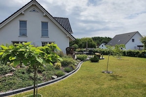 Um bairro residencial com uma casa branca, gramado bem cuidado e um jardim com diversas plantas.