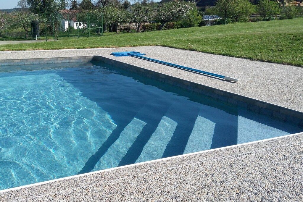 A swimming pool with clear water, surrounded by a pebble deck and a grassy area.